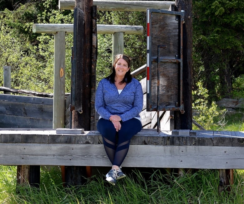 A woman wearing a blue top poses on a wooden structure.