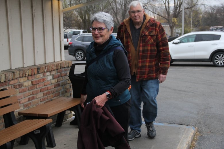 A woman in a light green jacket walk on a sidewalk as her husband trails behind her.