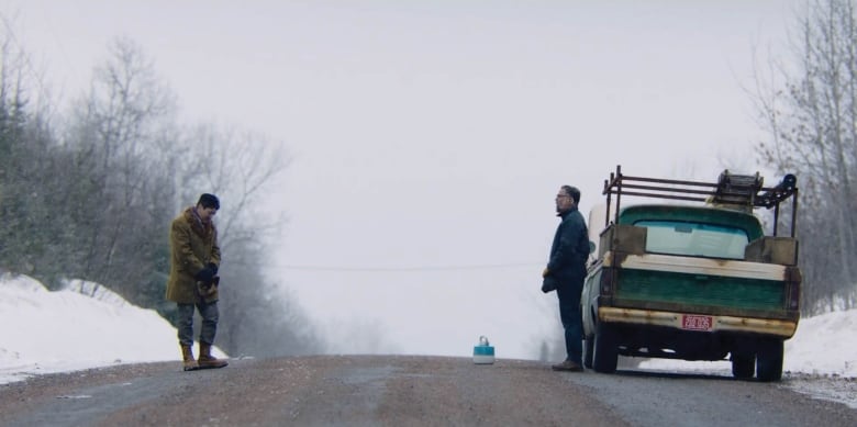 Two men standing across from each other on an empty road. The man on the right is standing in front of a beat-up pickup truck.