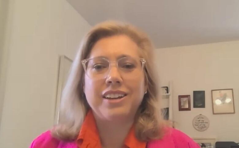 A blond-haired woman wearing a pink and orange shirt and clear-framed glasses speaks.