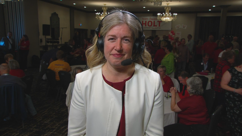 A blond woman in a white blazer, wearing a headset, closed-mouth smiles.