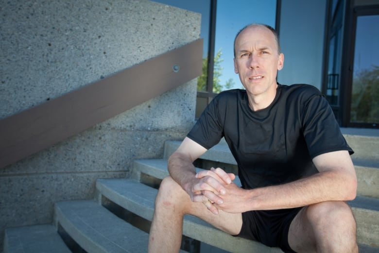 A man wearing a black t-shirt and shorts sits on steps with his hands crossed.