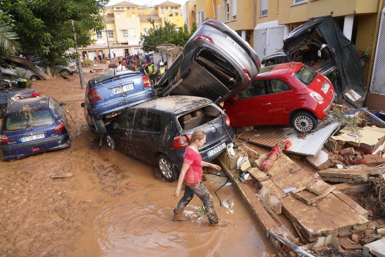 A mud-covered woman walks through brown water past overturned cars toward wood debris.