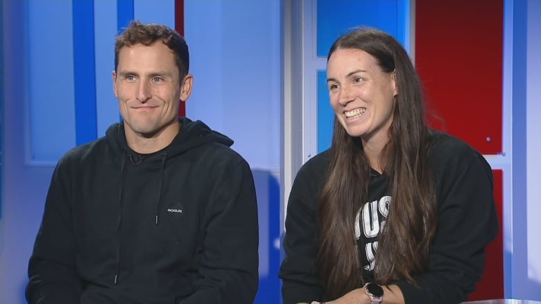 Man and woman in black hoodies smile as they sit on the CBC News: Compass anchor desk. 