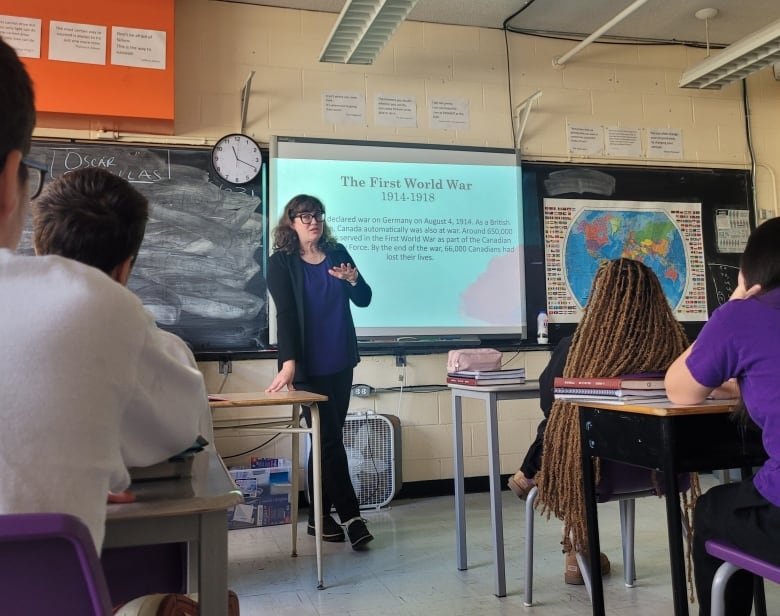 Students seated in a classroom are seen from behind listening to a female teacher, at centre, who stands by a projection screen showing a slide marked 'The First World War.' 