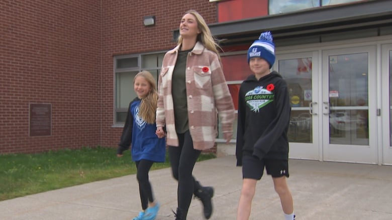 A mother in a pink checked shacket, flanked by a girl and a boy, walks away from a school