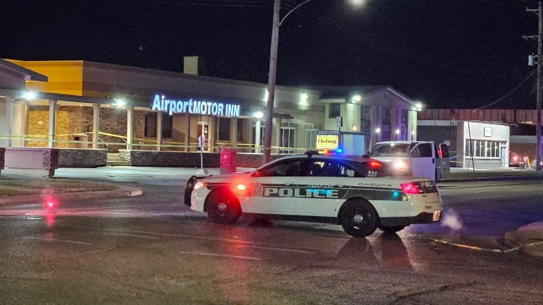 A police car is parked near a taped-off road. It is night time. The lights of a hotel can be seen in the background.