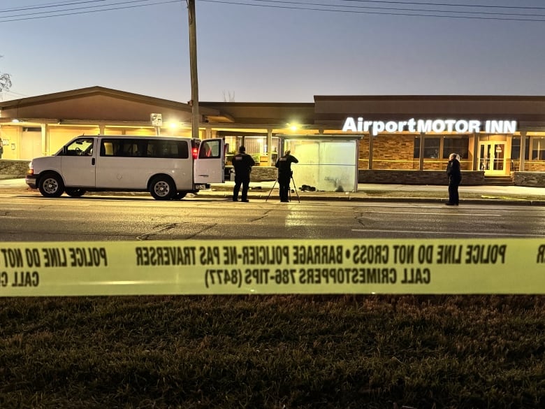 A band of yellow police tape is in the foreground.  Police officers in the background take photo at a frosted-up bus shelter in front of the Airport Motor Inn.