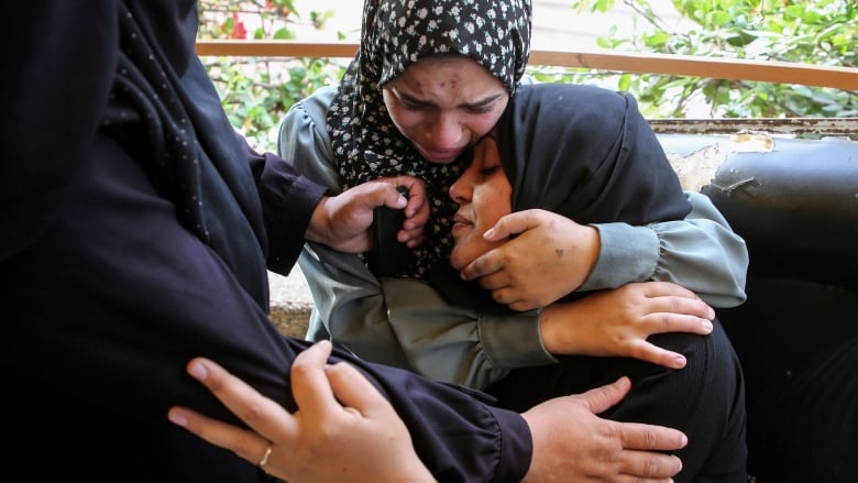 Two women in religious head coverings embrace while looking mournful in a closeup photo.