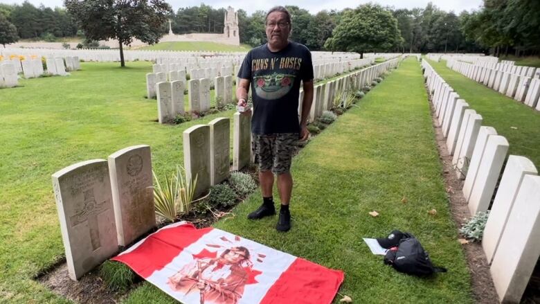 A man stands in a cemetery, near a Canadian flag on the ground with an image of an Indigenous man in traditional dress superimposed on the maple leaf.