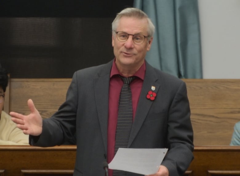 A grey-haired, bespectacled man in a gray suit and tie and maroon shirt with a poppy on his lapel stands with a paper in his hand in the P.E.I. legislature. 