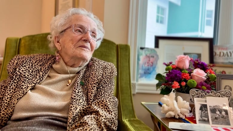 An elderly woman sits in a chair besides a floral arrangement and some photos on a table.