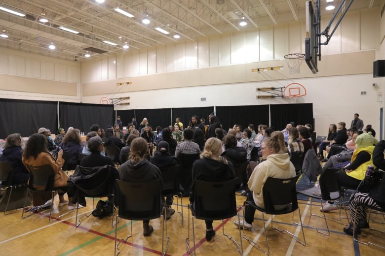 Students stand in the centre of concentric circles of people sitting in chairs in a gymnasium.