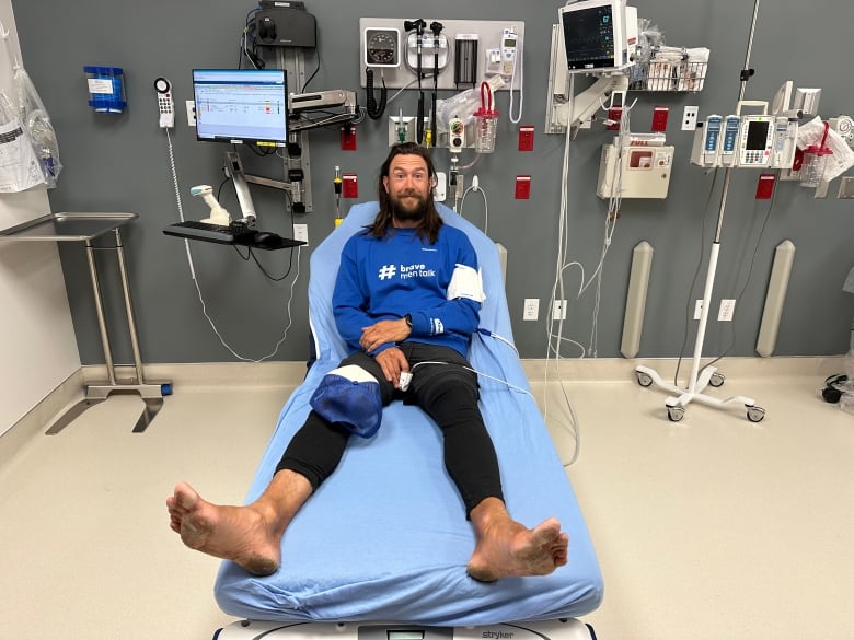 A mustachioed man with long hair and extremely dirty feet smiles as he lays in a hospital bed. 