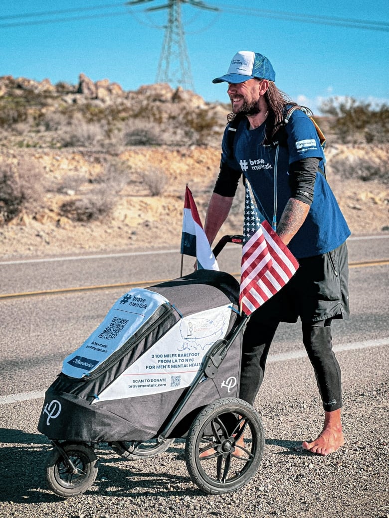 A mustachioed man with long hair and a trucker hat walks down a highway barefoot, pushing a car with an American and a Dutch flag.