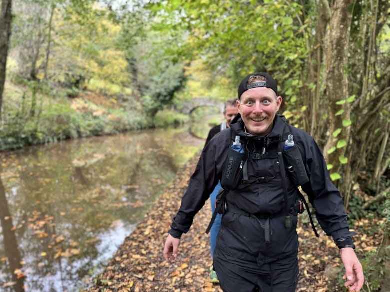 A smiling mustachioed man walks along a creek