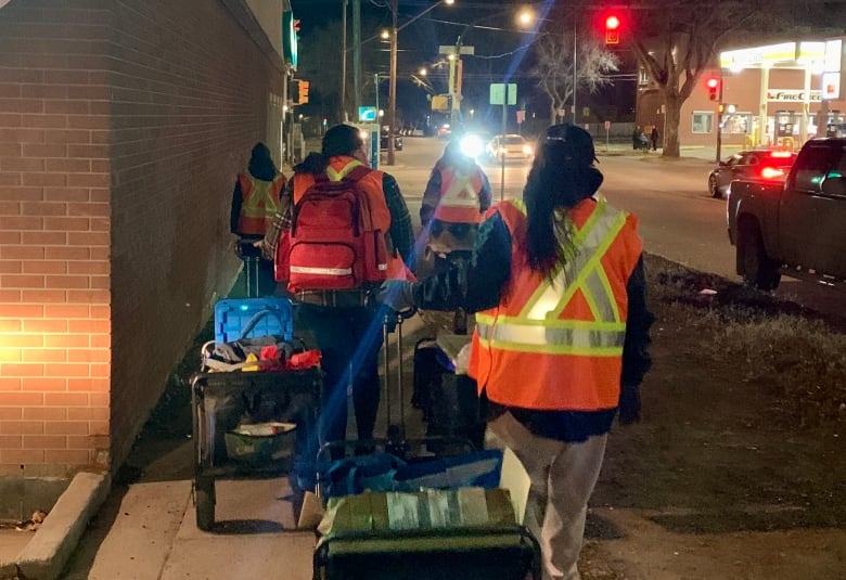 A group of people in orange safety vests pulling carts walking on a sidewalk at night.