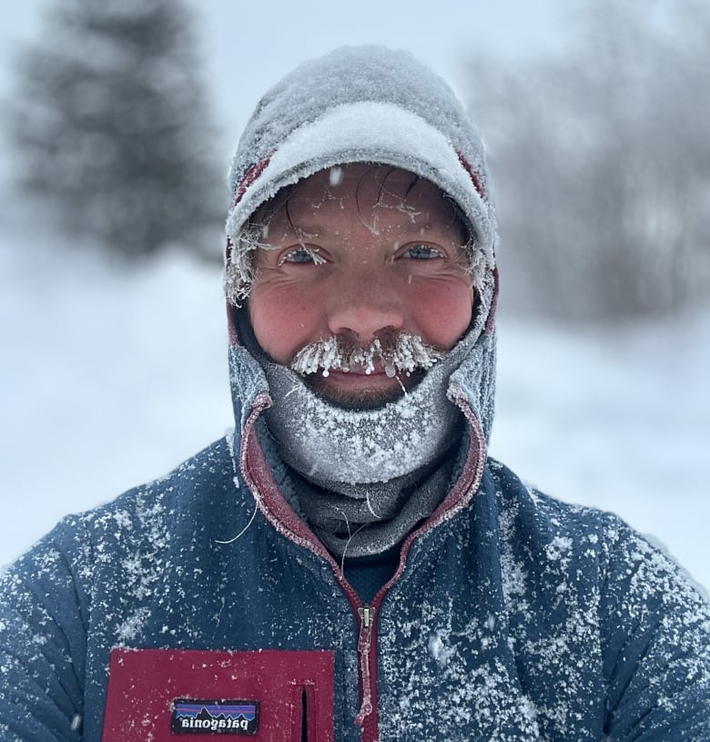 A mustached man smiles at the camera. He is covered in snow and ice. 