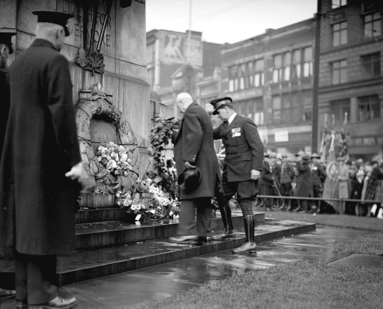 Two men, one in a military uniform, lay a wreath at the Victory Square Cenotaph while a crowd looks on. The photo is quite old.