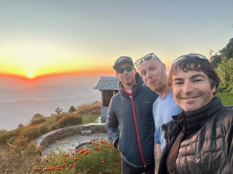 Three men pose in a row with a sunset behind them.