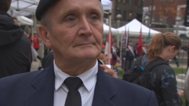 A man in a suit looks toward the camera with flags and a crowd in the background.