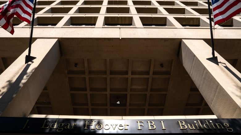 An exterior shot of a imposing building flanked by American flags, with the words 'J. Edgar Hoover FBI Building' on a sign.