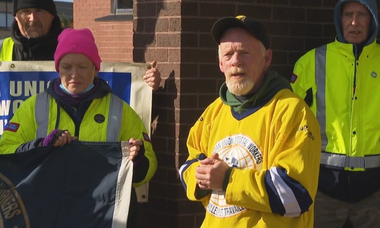 Man in baseball hat and yellow jersey stands in front of crowd holding banners. 