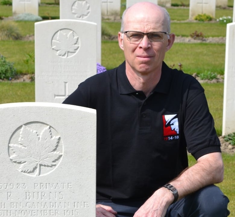 A man in glasses crouches next to a white military gravestone.