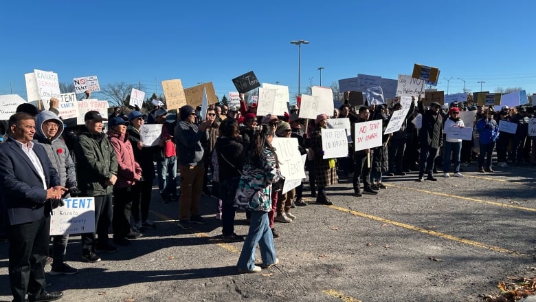 A large crowd of people, many of whom are carrying protest signs, gathers together in a parking lot on a sunny day.