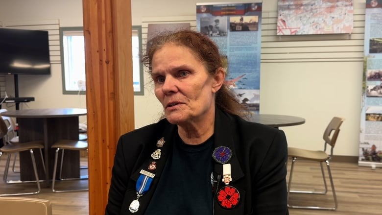 A light-skinned woman with amber-brown hair is sitting in a meeting room. She is wearing a dark shirt and black jacket, with medals, pins and poppies on her lapel.