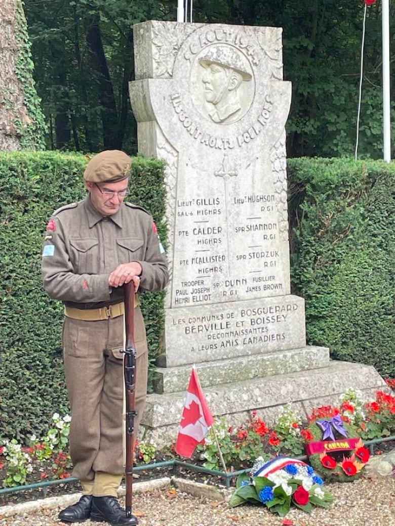 A soldier, wearing a military uniform, bows his head while standing beside a stone memorial.