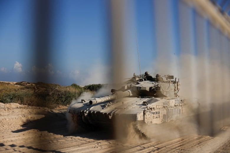 A tank drives down a dusty path, visible through the blurred bars of some sort of fence. 