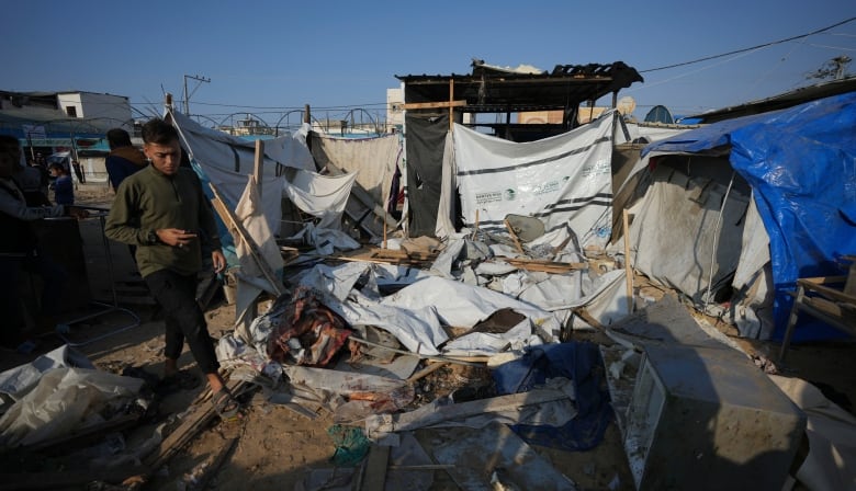 A person walks through a destroyed area, where fabric and tarps lie strewn on the ground with pieces of wood and rubble. Makeshift shelters are visible in the background. 