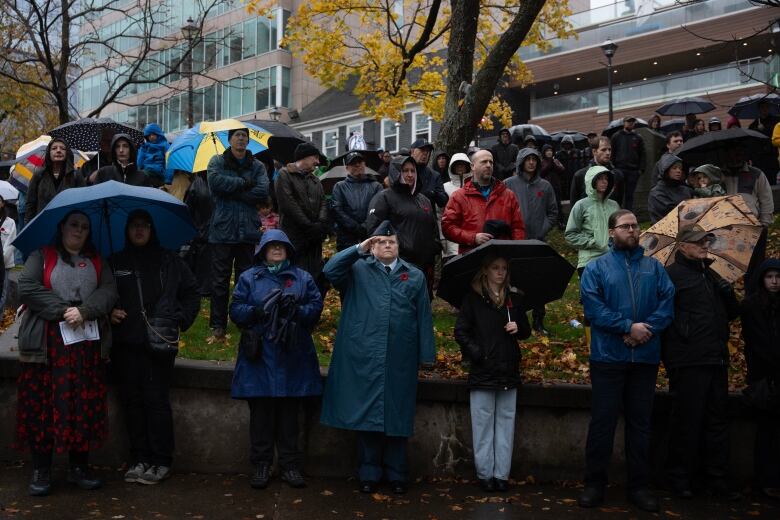 A group of people standing in the rain at a Remembrance Day ceremony and one person in a beret saluting. 