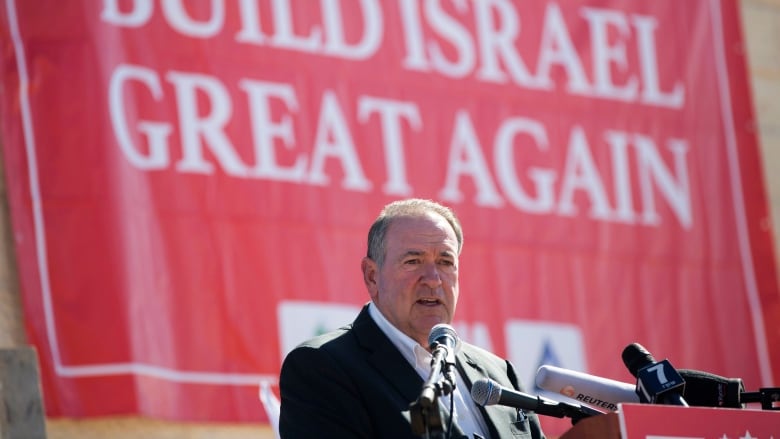 A man in a suit stands at a podium. A red banner behind him says 