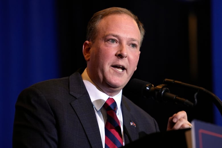 A cleanshaven man wearing a suit and tie speaks while standing at a podium.