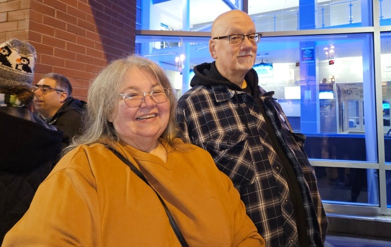 A woman with glasses and long grey hair stands beside a tall bald man who also wears glasses.