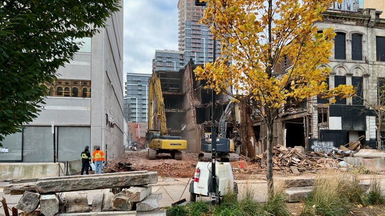 Two workers watch two excavators working on the site of a building which has been partially demolished in a downtown city block.