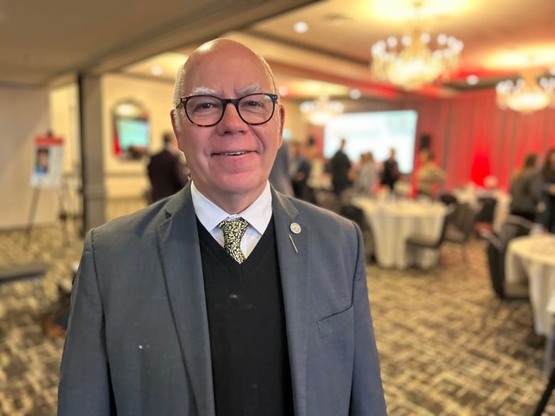 A man with black rimmed glasses and a suit jacket and tie smiles at the camera with people standing around next to tables behind him. 