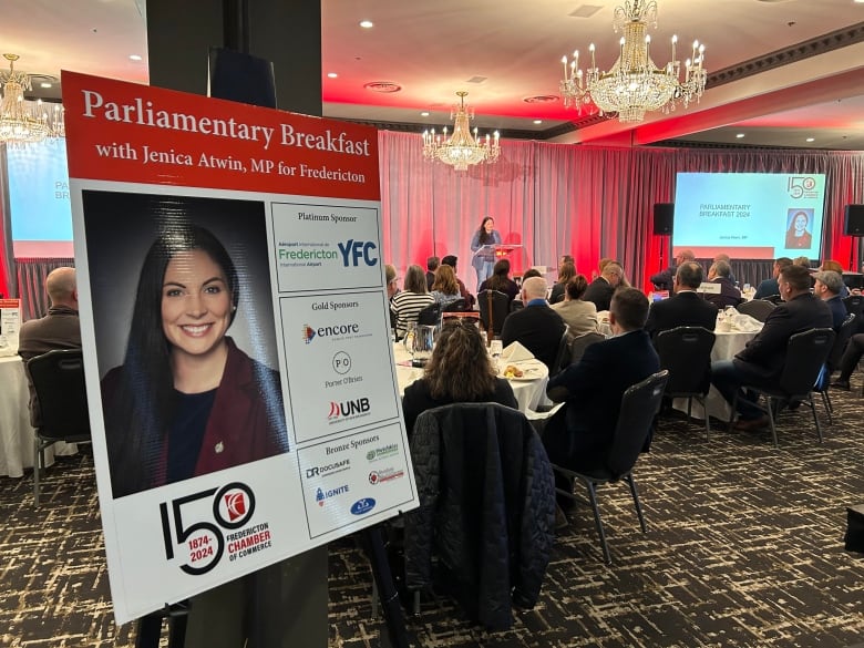 A woman with brown hair and a blue blazer stands at a podium addressing a crowd of people sitting at tables, with a poster of her in the foreground. 