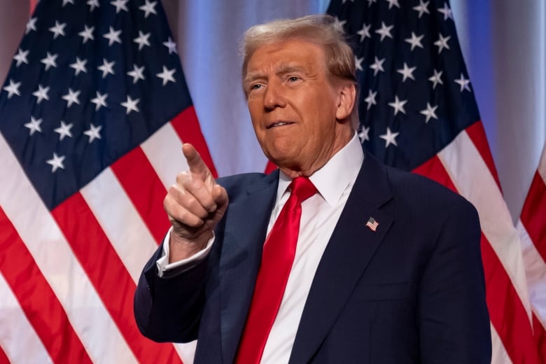 A man in a blue suit and a red tie points as he stands on a stage in front of American flags.