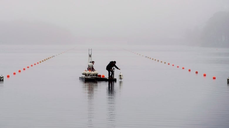 A foggy lake scene shows a person in dark clothing standing on a floating platform, leaning over the water to drop in a white buoy