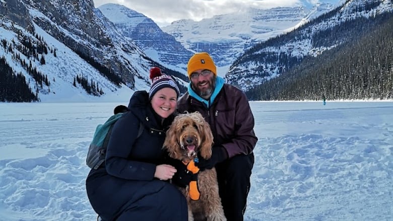 A man and woman crouch on ice with their dog, mountains in the background.