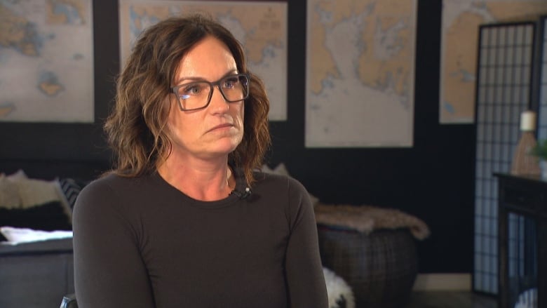 A woman with brown hair and glasses looks off to the side. She wears a black long-sleeved shirt. The wall behind her is covered in posters of world maps. 