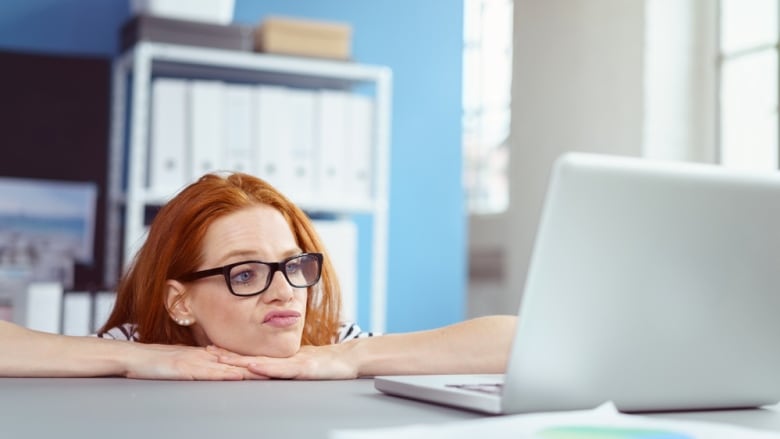 Frowning red haired female worker in eyeglasses with bored expression with hands under chin at desk behind laptop computer