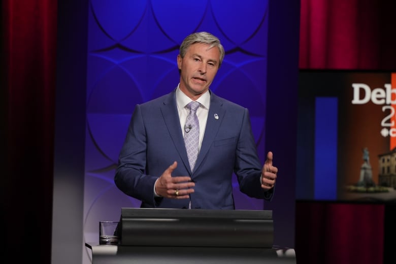 A man with short, grey hair gestures while speaking behind a podium. He wears a navy suit with a violet tie. He stands in front of a background illuminated blue.