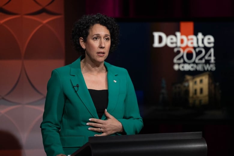 A woman with dark, curly hair and wearing a green blazer speaks behind a podium. 