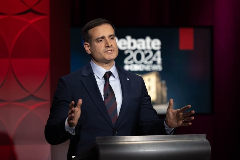 A man with short, dark hair speaks and gestures with his hands behind a podium.