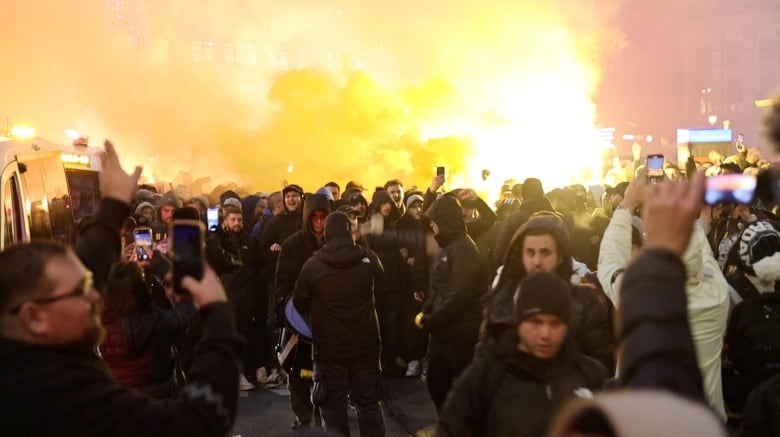 A group of men, some holding cellphones, demonstrate and light flares.