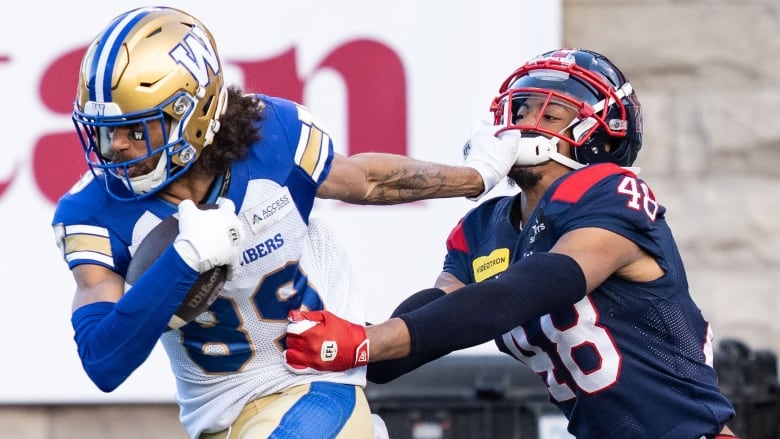 A player in white holds the ball in his right hand while reaching back with his left hand to push the face of an opponent in blue who is holding onto his jersey.